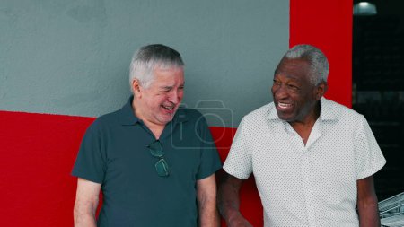 Foto de Joyful Interaction of Two Diverse Male Senior Friends, Smiling and Laughing Together, Leaning on Sidewalk Wall (en inglés). Auténticos ancianos felices de la vida real - Imagen libre de derechos
