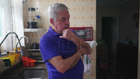 Photo for One pensive senior man in deep mental reflection standing in kitchen at home, pondering decision with hand in chin - Royalty Free Image