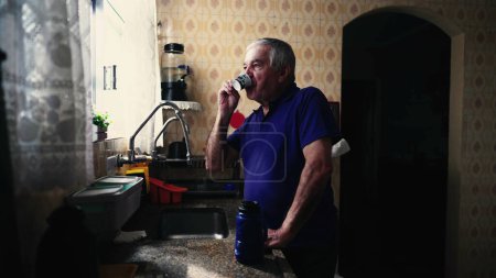Téléchargez les photos : Homme âgé dégustant un café avec un regard contemplatif regardant par la fenêtre de la maison le matin calme, personne âgée par évier sirotant une boisson chaude - en image libre de droit