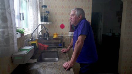 Photo for One pensive elderly man standing by kitchen sink staring out by window. Domestic lifestyle scene of a contemplative older male person in 70s pondering life - Royalty Free Image