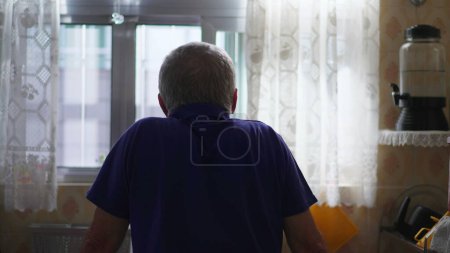 Photo for Back of contemplative senior man standing by kitchen sink staring outside through window view. Pensive eldery person in mental reflection at home alone - Royalty Free Image