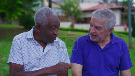 Photo for Two older diverse friends conversing at park bench. A retired African American and a caucasian senior people talking outside - Royalty Free Image