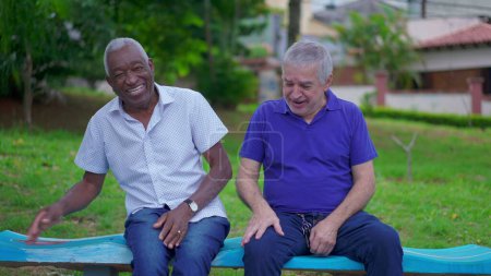 Photo for Two diverse senior talking and laughing sitting on park bench in Brazil. Candid authentic people real life laugh and smile in old age retirement golden years - Royalty Free Image