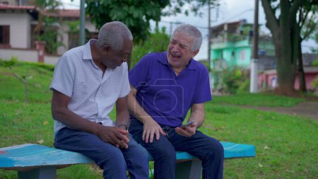 Photo for Two diverse seniors looking at phone sitting on park bench outdoors laughing and smiling together. Older friend sharing online media content to companion depicting authentic friendship - Royalty Free Image