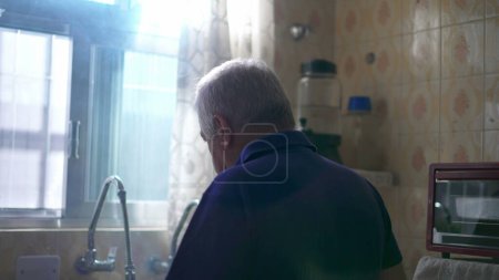 Foto de Espalda de hombre de edad avanzada pensativo de pie junto a la ventana de la cocina mirando hacia fuera con la luz del sol brilla a través de. La soledad contemplativa la emoción del anciano en la vejez - Imagen libre de derechos
