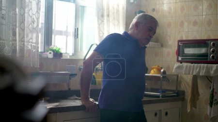 Téléchargez les photos : Pensif homme retraité debout près de la fenêtre de l'évier de cuisine contemplant la vie dans la solitude à la maison avec brume - en image libre de droit