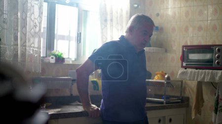 Foto de Pensativo anciano jubilado de pie junto a la ventana del fregadero de la cocina contemplando la vida en soledad en casa con neblina - Imagen libre de derechos