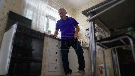 Photo for Pensive Senior man standing by kitchen sink at home alone. Low-angle domestic scene of One contemplative elderly person in solitude - Royalty Free Image