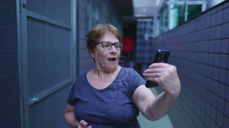 Foto de Mujer mayor reaccionando con SHOCK al contenido del dispositivo de teléfono celular, señora anciana mirando la pantalla del teléfono inteligente con incredulidad, de pie en la residencia doméstica - Imagen libre de derechos