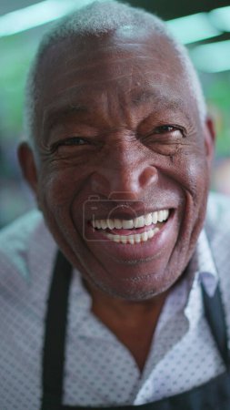 Téléchargez les photos : Portrait d'un heureux homme afro-américain âgé aux cheveux gris et aux rides, portant un tablier noir, souriant à la caméra avec une expression joyeuse. Charismatique et sympathique employé - en image libre de droit