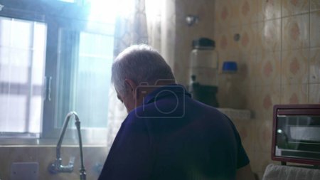 Téléchargez les photos : Le dos d'un vieil homme cher debout près de la fenêtre de la cuisine, regardant dehors avec la lumière du soleil brille à travers. Émotion contemplative de solitude des personnes âgées âgées - en image libre de droit