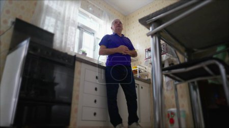 Téléchargez les photos : Pensive Senior homme debout près évier de cuisine à la maison seul. Scène domestique à faible angle d'une personne âgée contemplative dans la solitude - en image libre de droit