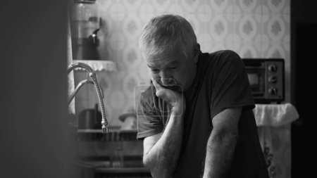 Photo for Senior man suffering from depression at home alone leaning by kitchen sink in dramatic black and white, monochromatic scene of person in despair succumbed by hardship - Royalty Free Image