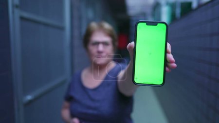 Téléchargez les photos : Femme âgée tenant dispositif de téléphone portable avec écran vert chroma clé sur l'affichage debout près du couloir balcon à la maison - en image libre de droit