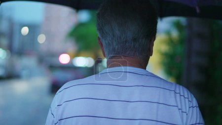 Téléchargez les photos : Vue arrière de l'homme âgé marchant dans la rue avec parapluie pendant la journée de pluie - en image libre de droit