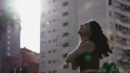 Téléchargez les photos : Joyeux jeune femme se retournant tout en se tenant dehors dans la lumière du soleil avec les mains dans la poitrine - en image libre de droit