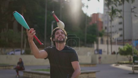 Téléchargez les photos : Homme Juggler artiste démontrant ses compétences à l'extérieur dans le parc urbain de la ville. Personnage artiste jongler avec des objets dans l'air - en image libre de droit