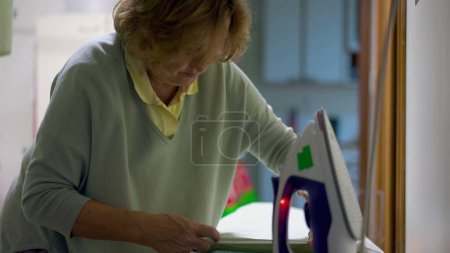 Photo for Ironing Out Domestic Chores_ Candid Footage of Senior Woman in Laundry Room. Authentic real life older lady doing everyday routine, ironing clothes with steam iron - Royalty Free Image