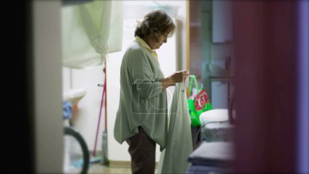 Photo for Efficient and Independent_ Senior Lady Tackles Household Chores in Her Laundry Room. Candid and authentic everyday domestic routine of older person folding sheets - Royalty Free Image