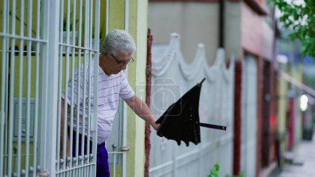 Foto de El hombre mayor que llega a casa mientras llueve, abre la puerta y cierra el paraguas. Senior caminando en la acera de la calle llega a la residencia - Imagen libre de derechos