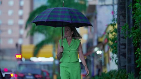 Téléchargez les photos : Femme face à la pluie pendant l'après-midi pluvieux dans la rue de la ville, personne contemplative marchant tenant parapluie - en image libre de droit