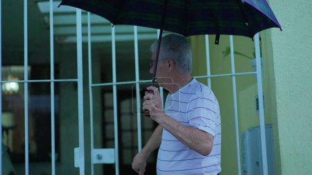 Téléchargez les photos : Un aîné ouvre la porte, sort de chez lui avec un parapluie sous la pluie, marche sur un trottoir urbain - en image libre de droit