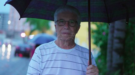 Foto de El hombre mayor camina hacia adelante sosteniendo el paraguas durante el día lluvioso. Una persona anciana caucásica madura paseando en la acera de la ciudad - Imagen libre de derechos
