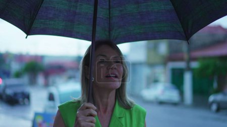 Téléchargez les photos : Femme joyeuse dans sa cinquantaine tenant parapluie, se promener dans la rue Rainy City - en image libre de droit