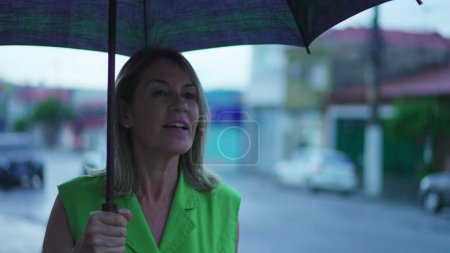 Téléchargez les photos : Femme joyeuse dans sa cinquantaine tenant parapluie, se promener dans la rue Rainy City - en image libre de droit