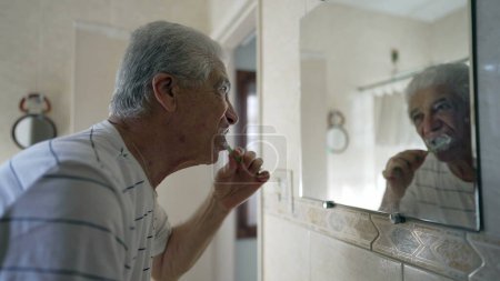 Photo for Senior man brushing teeth in front of bathroom mirror in casual domestic scene in old age morning routine. Elderly mature gray-hair person starting the day - Royalty Free Image