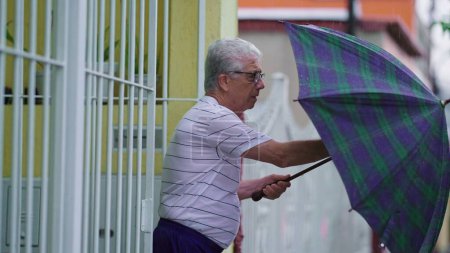 Téléchargez les photos : Homme âgé arrivant à la maison pendant la journée de pluie, tenant un parapluie et ouvrant la porte - en image libre de droit