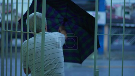 Téléchargez les photos : Homme mûr âgé se préparant pour une promenade dans la soirée pluvieuse, ouverture parapluie sur le trottoir de la rue - en image libre de droit