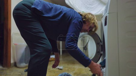 Téléchargez les photos : Une femme âgée met des vêtements à l'intérieur de la machine à laver. Scène domestique de la personne faisant ménage chrores routine d'activité - en image libre de droit