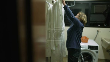 Téléchargez les photos : Candid senior woman putting sheets to dry at laudry room rack. Personne séchant des vêtements - en image libre de droit
