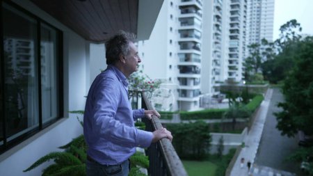 Photo for P''pOne older man standing at apartment balcony in the evening. Contemplative senior male person in 70s looking out - Royalty Free Image