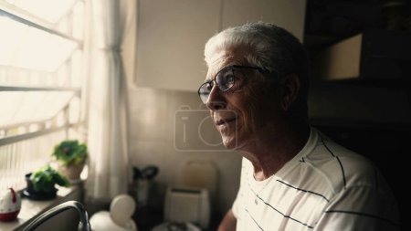 Téléchargez les photos : Homme plus âgé contemplatif regardant par la fenêtre dans la cuisine. Regard méditatif réfléchi d'un homme mûr réfléchissant sur la vie - en image libre de droit