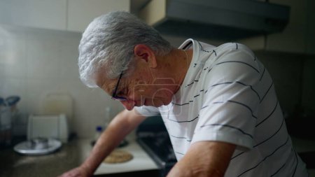 Téléchargez les photos : Homme mûr debout au comptoir de la cuisine aux prises avec la vieillesse et le regret. Homme âgé inquiet regardant vers le bas gelé dans la courtoisie - en image libre de droit
