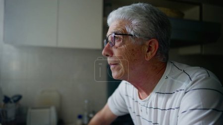 Téléchargez les photos : Homme mûr debout au comptoir de la cuisine aux prises avec la vieillesse et le regret. Homme âgé inquiet regardant vers le bas gelé dans la courtoisie - en image libre de droit