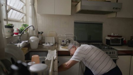 Foto de Hombre mayor de pie en fregadero de cocina organizando ollas y sartenes por ventana en casa. Auténtica escena doméstica de los jubilados de más edad - Imagen libre de derechos