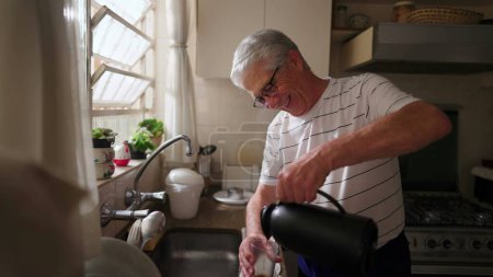 Photo for Mature man serving coffee with thermos bottle into glass by kitchen sink. Authentic domestic scene of retired older person starting the day ritual morning - Royalty Free Image