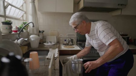 Foto de Hombre mayor de pie en fregadero de cocina organizando ollas y sartenes por ventana en casa. Auténtica escena doméstica de los jubilados de más edad - Imagen libre de derechos