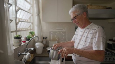 Foto de Feliz hombre mayor secando platos junto al fregadero de la cocina. Auténtico estilo de vida escena doméstica de la persona mayor madura en el ritual casero - Imagen libre de derechos
