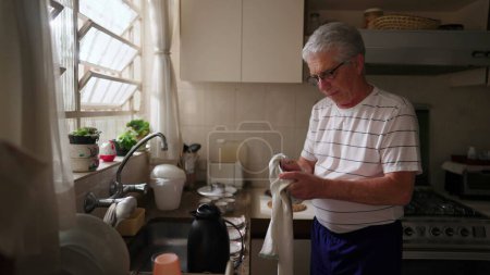 Téléchargez les photos : Homme âgé à la retraite séchant des ustensiles de cuisine debout à l'évier. Scène de style de vie domestique de la personne âgée faisant des routines à la maison - en image libre de droit