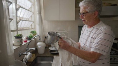 Happy senior man drying dishes by the kitchen sink. Authentic domestic scene lifestyle of mature older person at home ritual