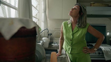 Téléchargez les photos : Une femme d'âge moyen anxieuse debout dans la cuisine souffrant de stress et de détresse. Une femme caucasienne dans les années 50 éprouvant regret et anxiété - en image libre de droit
