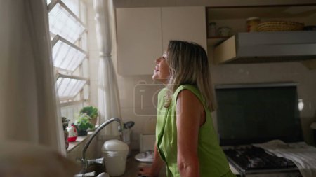 Foto de Una alegre mujer mayor de mediana edad de pie junto a la ventana en la cocina mirando hacia fuera con expresión feliz. Una persona femenina de unos 50 años con una emoción feliz y pensativa - Imagen libre de derechos