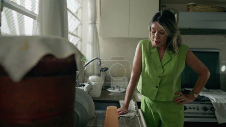 Foto de Una mujer ansiosa de mediana edad de pie en la cocina sufriendo de estrés y angustia. Una mujer caucásica en los años 50 sintiendo pesar y ansiedad - Imagen libre de derechos