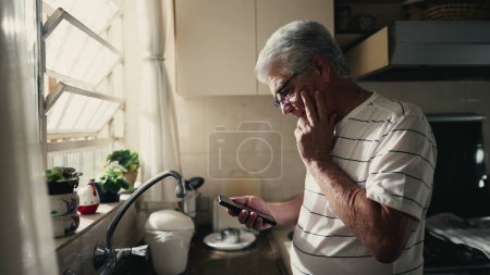 Foto de Hombre de edad avanzada pensativo mirando la pantalla del teléfono ponderando dilema y la toma de decisiones con un dispositivo de teléfono inteligente mientras está de pie en la cocina del hogar por la ventana - Imagen libre de derechos