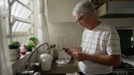 Foto de Un hombre mayor mirando el dispositivo de teléfono móvil mientras está de pie en la cocina casera por la ventana. Pensativo mayor jubilado hombre caucásico utilizando el dispositivo de tecnología moderna - Imagen libre de derechos