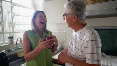 Foto de Pareja mayor mirando el dispositivo de teléfono celular juntos en la cocina. Esposa mostrando pantalla a marido, auténtica vida real risa y sonrisa - Imagen libre de derechos